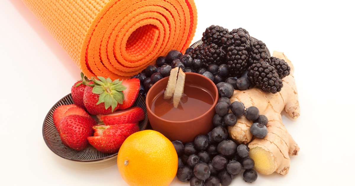 Bright orange yoga mat with ginger tea in a distinctive cup surrounded by bright berries, ginger root, and citrus. On white background.