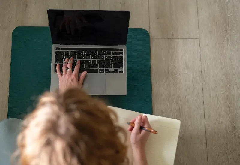 Overhead view of a student taking notes at a laptop, representing the online learning portion of Mount Madonna's hybrid YTT 300
